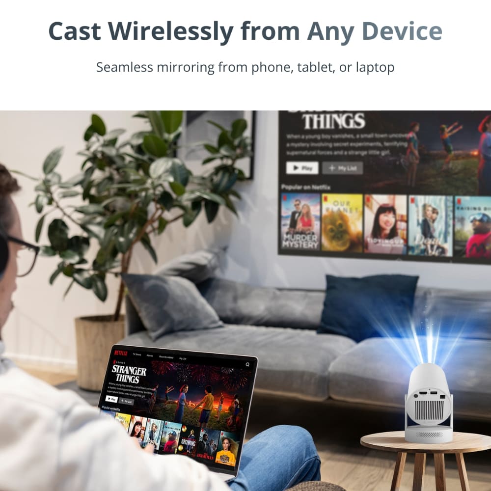 A man wearing headphones casts Netflix's Stranger Things from a laptop onto a living room wall using a white portable projector emitting a blue light beam showing other Netflix titles like Murder Mystery and Our Planet