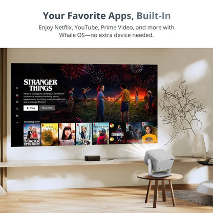 A white projector on a small wooden table projects a large Netflix interface featuring Stranger Things with colorful fireworks and a trending shows row onto a wall in a minimalist living room
