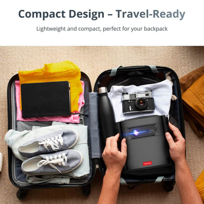A pair of hands placing a compact black projector emitting blue light into a suitcase filled with travel items including a vintage silver camera, grey sneakers, and colorful clothing