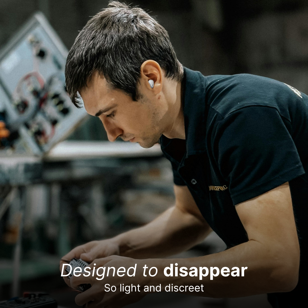 Photograph of a man wearing a discreet white wireless earbud in his right ear, intently focused on intricate wiring in a blurred industrial workshop while wearing a dark polo shirt