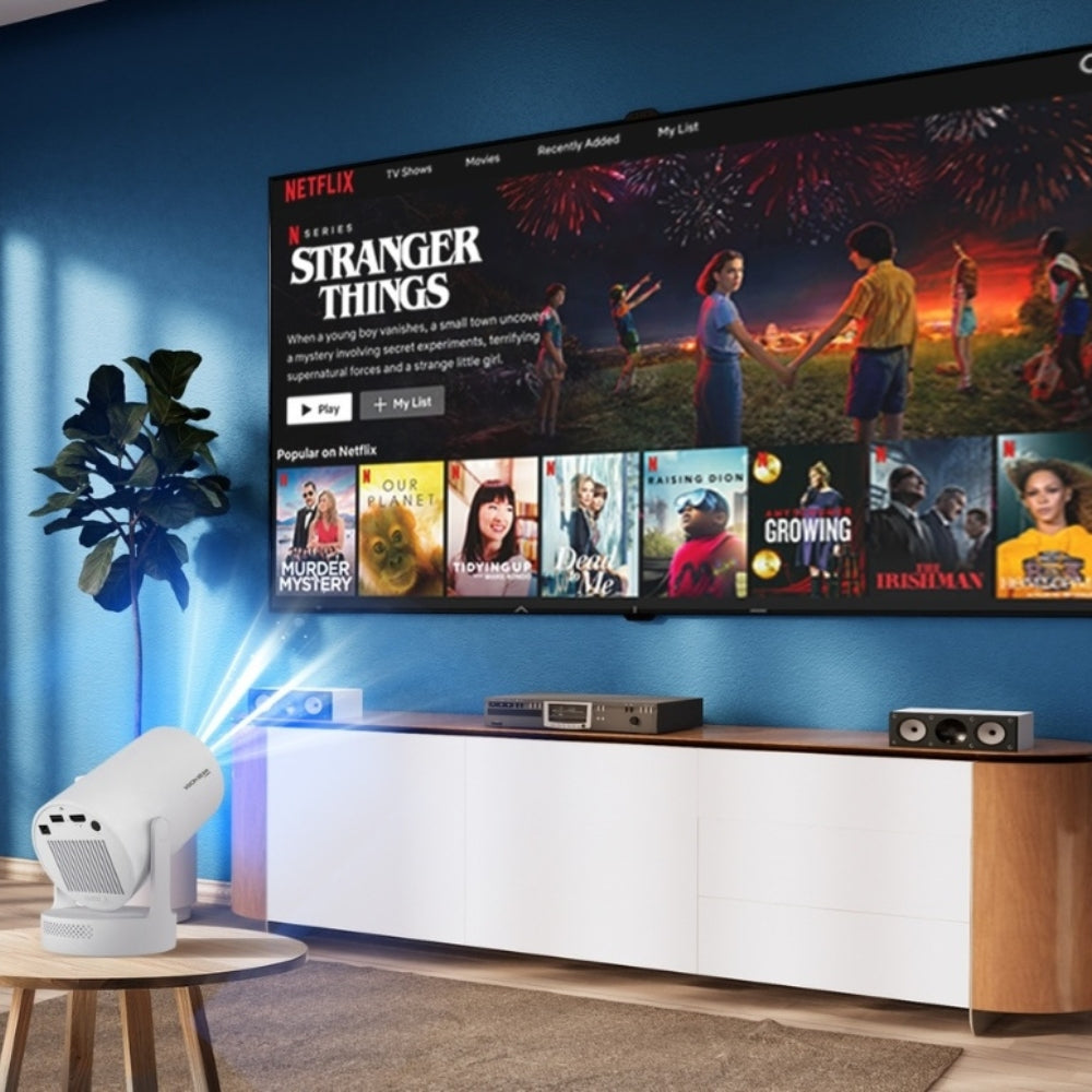 Photograph of a white projector casting a blue light beam onto a blue wall where a large smart TV displays Netflix's Stranger Things with colorful fireworks, positioned above a modern white and wood media console in a contemporary living room