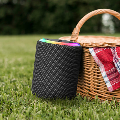 Black portable speaker with a rainbow LED ring next to a woven wicker picnic basket featuring a red plaid cloth on green grass