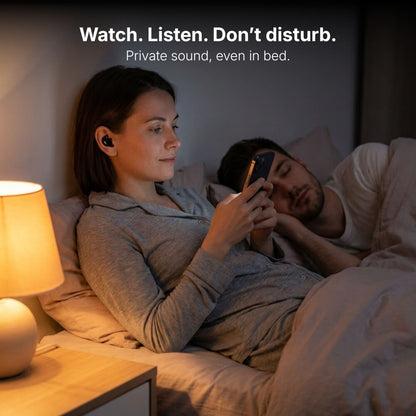 Photograph of a woman in grey pajamas wearing a black wireless earbud, focused on her glowing phone screen while a man sleeps peacefully beside her in a dimly lit bedroom, with a warm bedside lamp