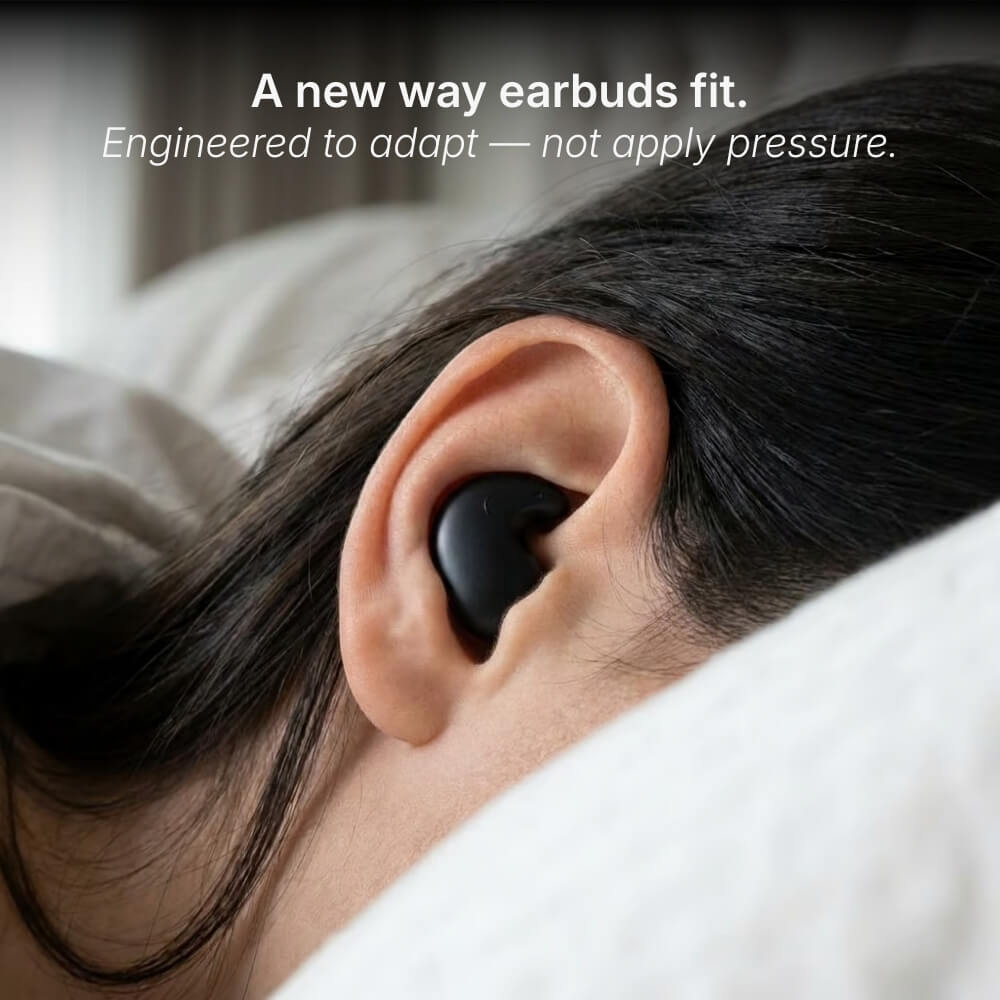Digital photograph showing a close-up of a person's ear with a sleek black earbud nestled comfortably, lying on soft white bedding, overlaid with the promotional text "A new way earbuds fit Engineered to adapt — not apply pressure"
