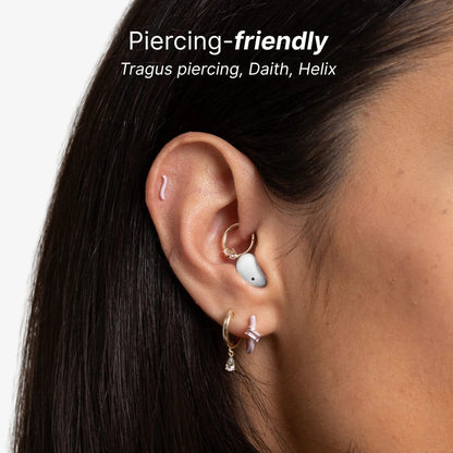 A close-up image of a woman's ear with text Piercing-friendly Tragus piercing Daith Helix showing a white earbud a gold daith hoop with clear gems a pink wave helix stud and two lobe piercings with gold and lilac enamel hoops