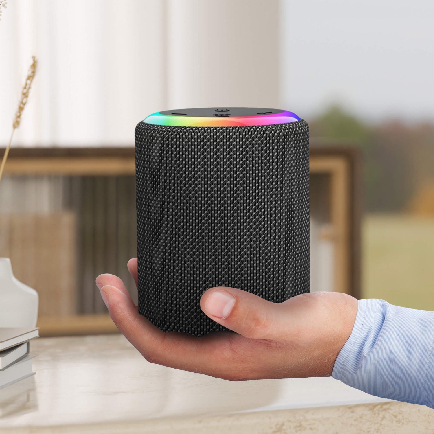 A black textured smart speaker with a vibrant rainbow LED light ring is held by a hand wearing a blue striped shirt, presented against a blurred indoor setting