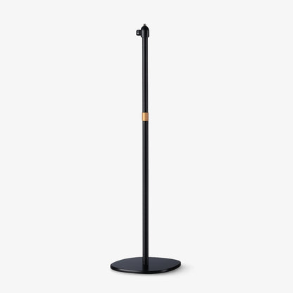 Product photograph of a minimalist matte black floor stand featuring a slim cylindrical pole, a prominent copper accent band, and a sleek oval base with an adjustable top mount