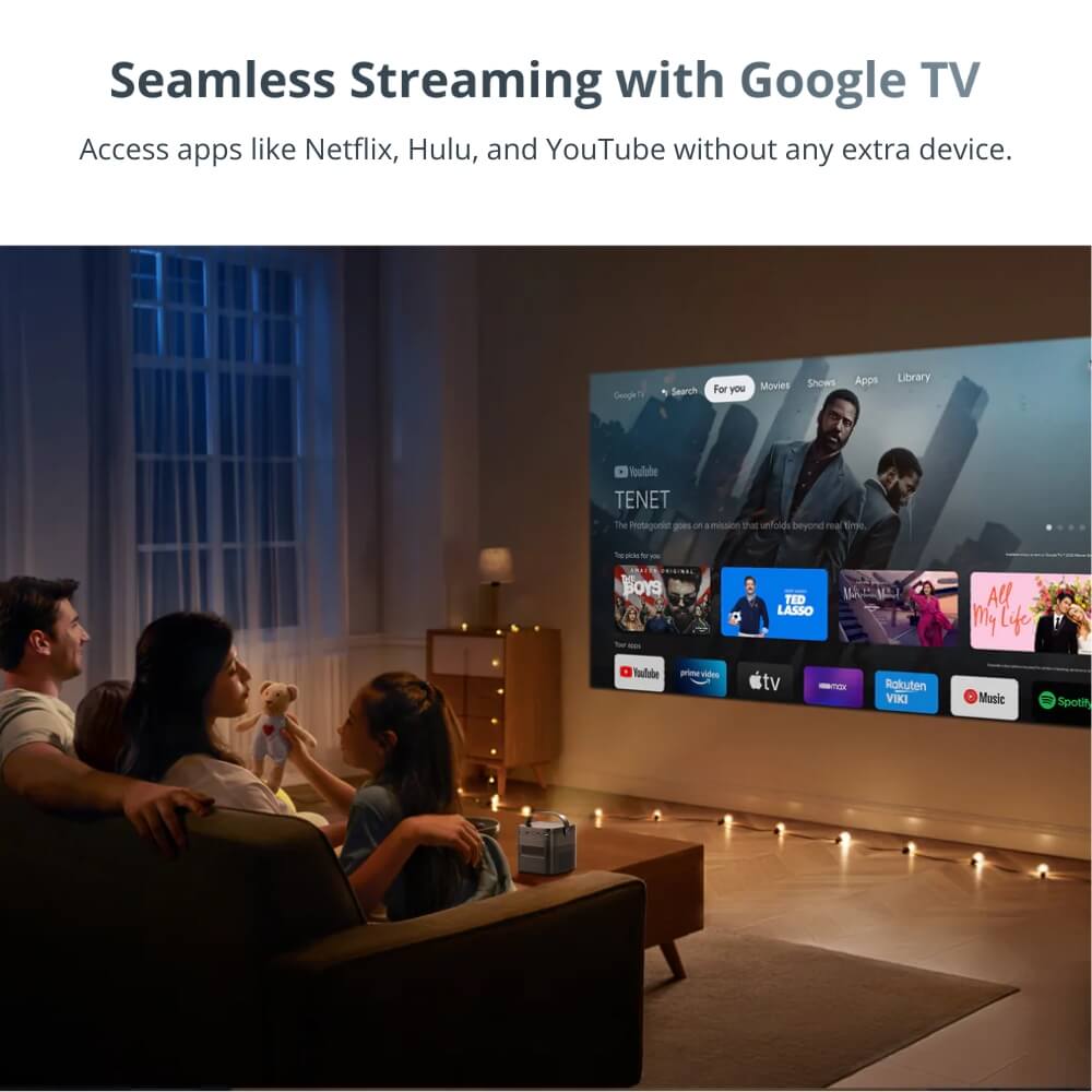 A family including a child holding a teddy bear relaxes on a couch in a dimly lit living room watching a large projection of the Google TV interface displaying titles like TENET and various streaming service apps with a small white projector on the coffee table and festive string lights on the floor
