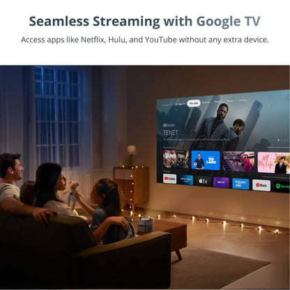 A family including a child holding a teddy bear relaxes on a couch in a dimly lit living room watching a large projection of the Google TV interface displaying titles like TENET and various streaming service apps with a small white projector on the coffee table and festive string lights on the floor